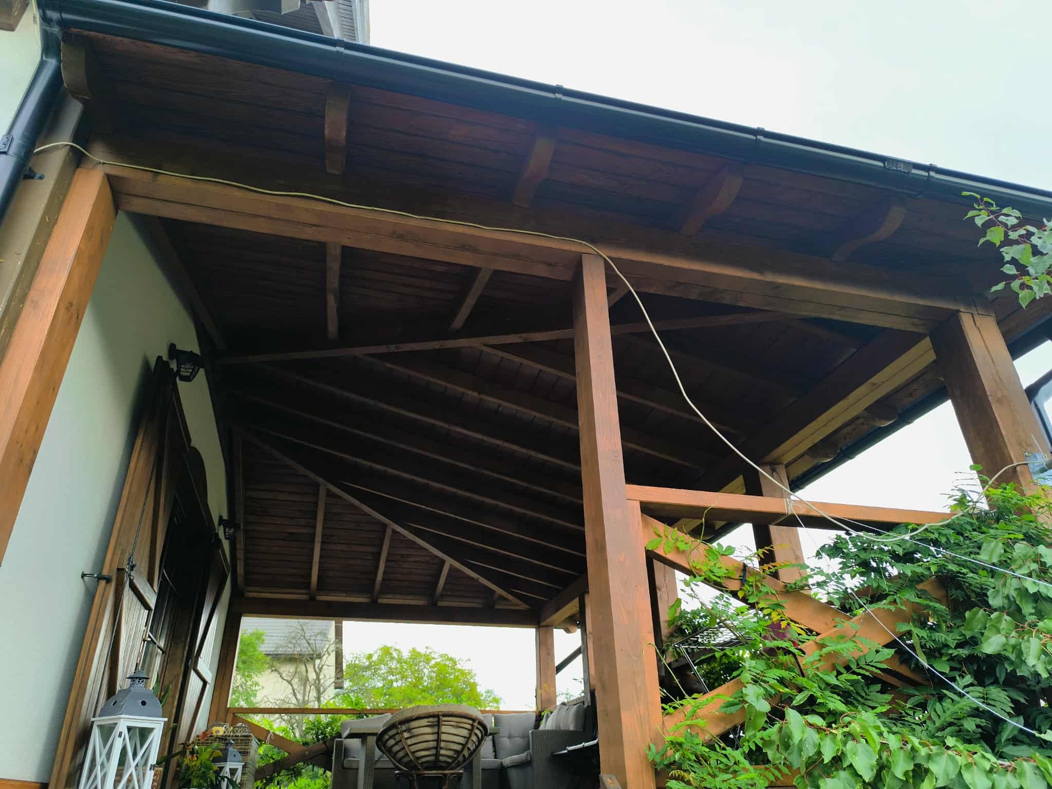 Zadaszona, drewniana konstrukcja z widocznym fragmentem dachu i zielenią. Widok z dołu ukazuje detale belek i konstrukcji nośnej. Elementy wyposażenia w tle.