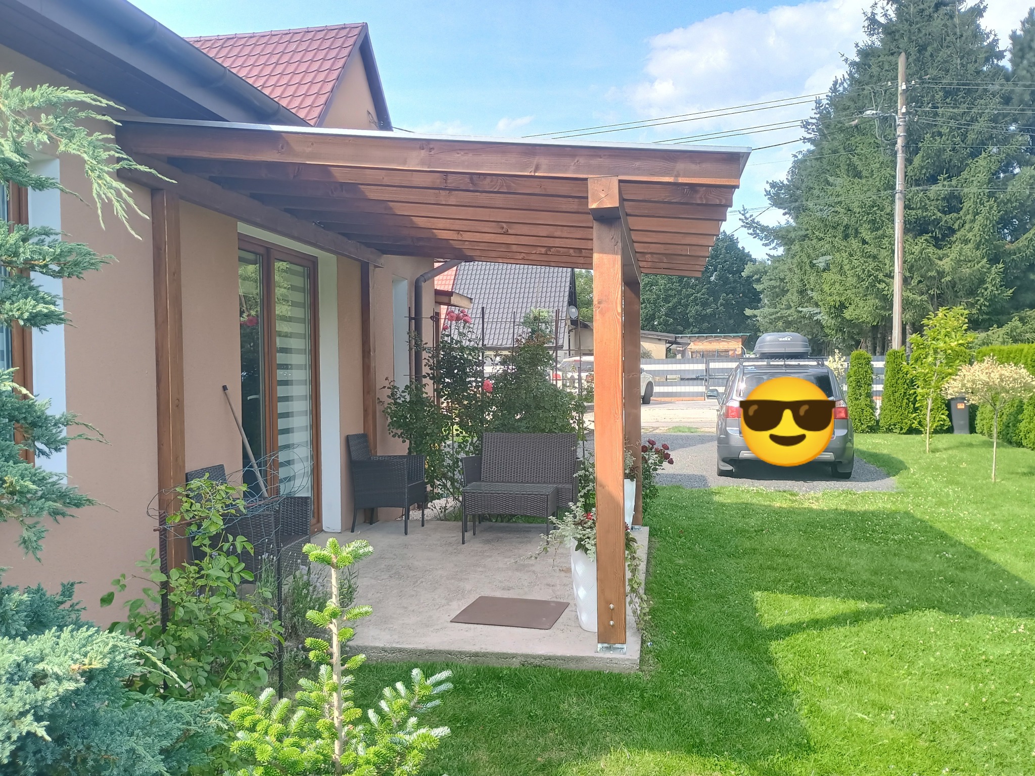 Drewniana pergola przy domu z meblami ogrodowymi i roślinami w donicach. Widok na zadbany trawnik i samochód w tle. Jasne, słoneczne oświetlenie.