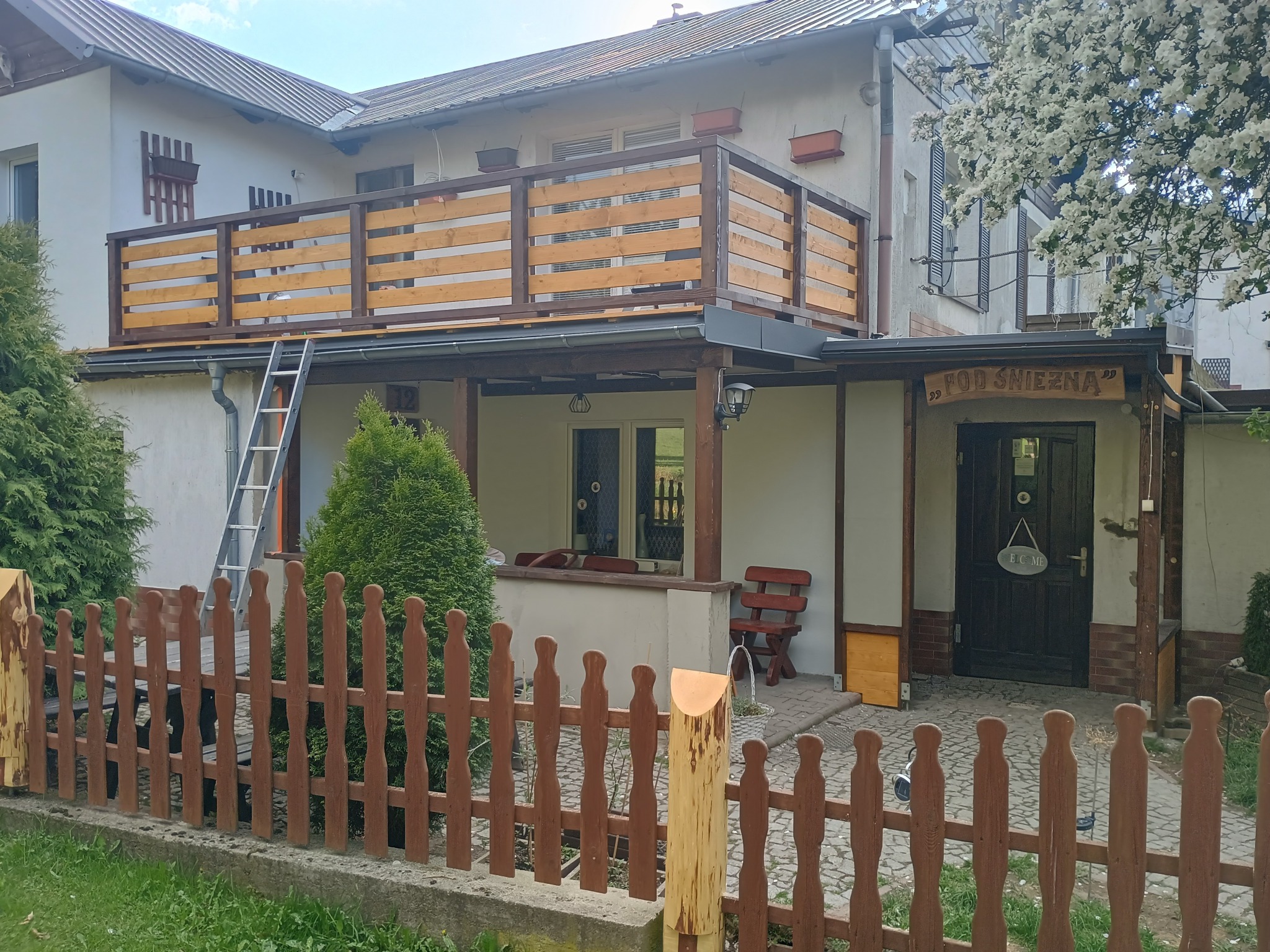 Dwukondygnacyjny dom z drewnianą balustradą na balkonie i ogrodzeniem z palików. Drabina oparta o balkon. Widoczny fragment kwitnącego drzewa.