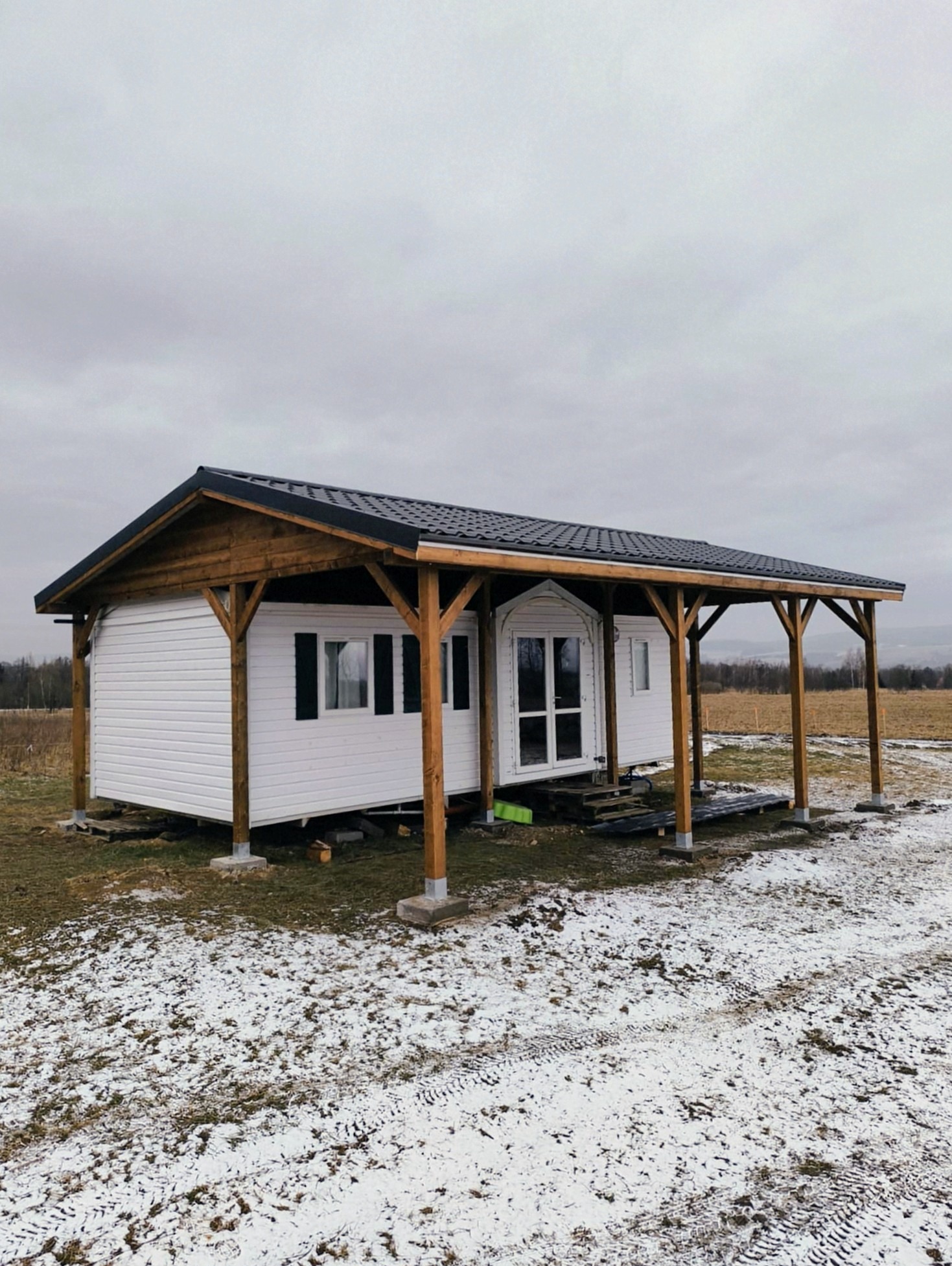 Biały dom szkieletowy z drewnianą konstrukcją dachu na betonowych podstawach, otoczony polem z resztkami śniegu pod pochmurnym niebem.