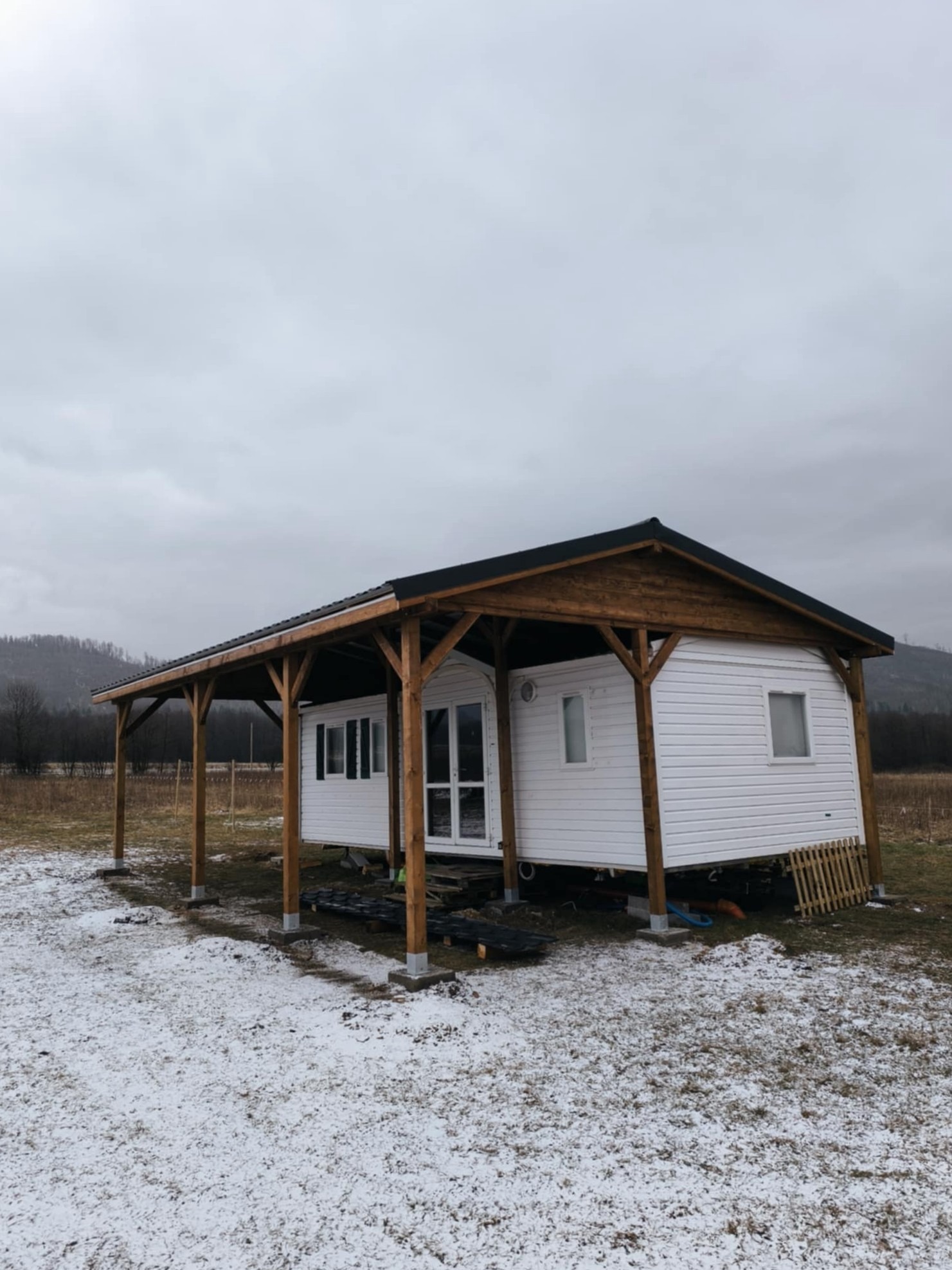 Biały dom mobilny z drewnianą konstrukcją zadaszenia, osadzony na betonowych fundamentach, na tle zimowego krajobrazu z górami w oddali.