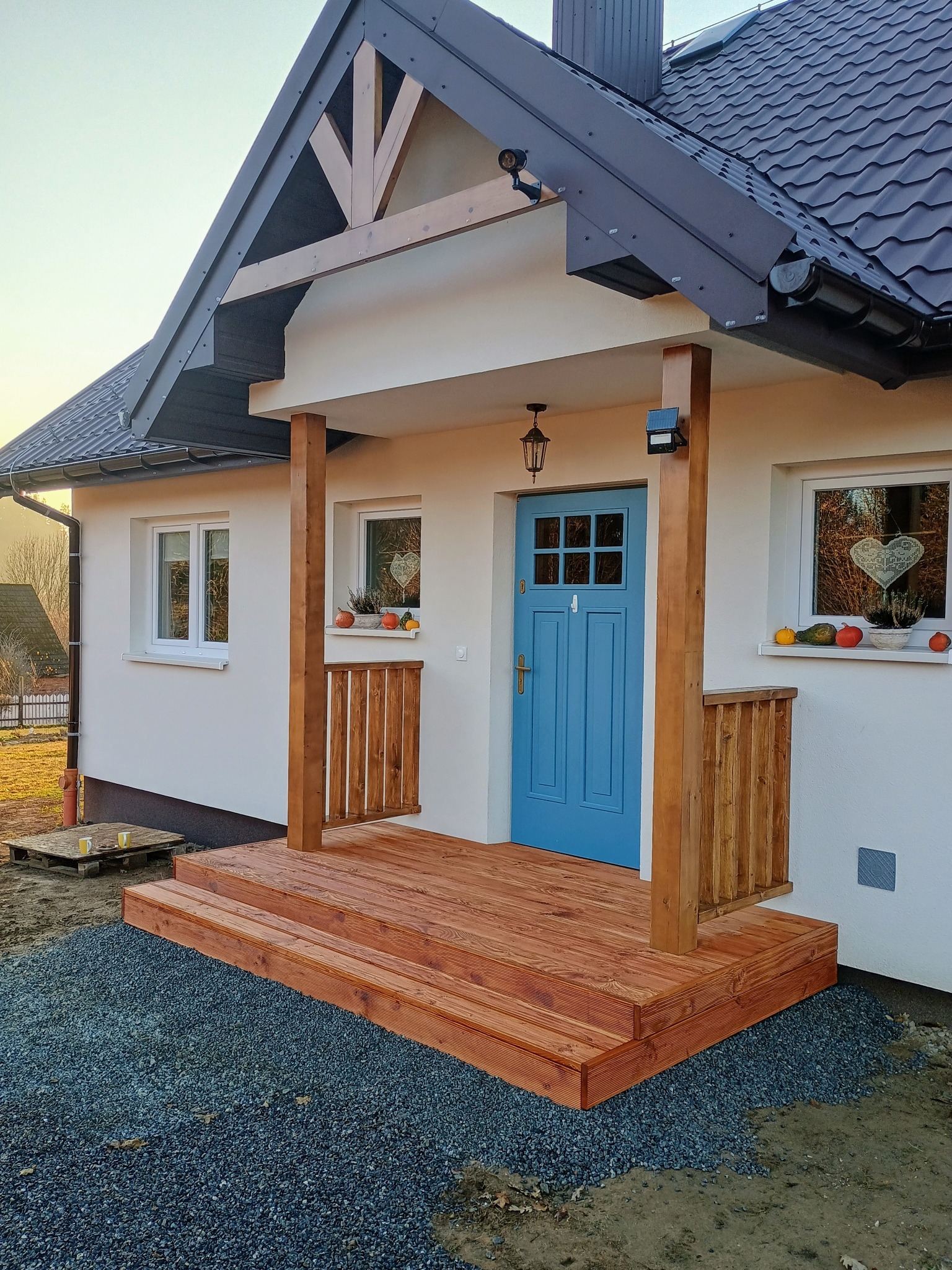 Nowoczesny dom z drewnianym tarasem wejściowym i balustradą. Błękitne drzwi wejściowe, dekoracje okienne. Ujęcie z poziomu gruntu, widoczne detale wykończenia.