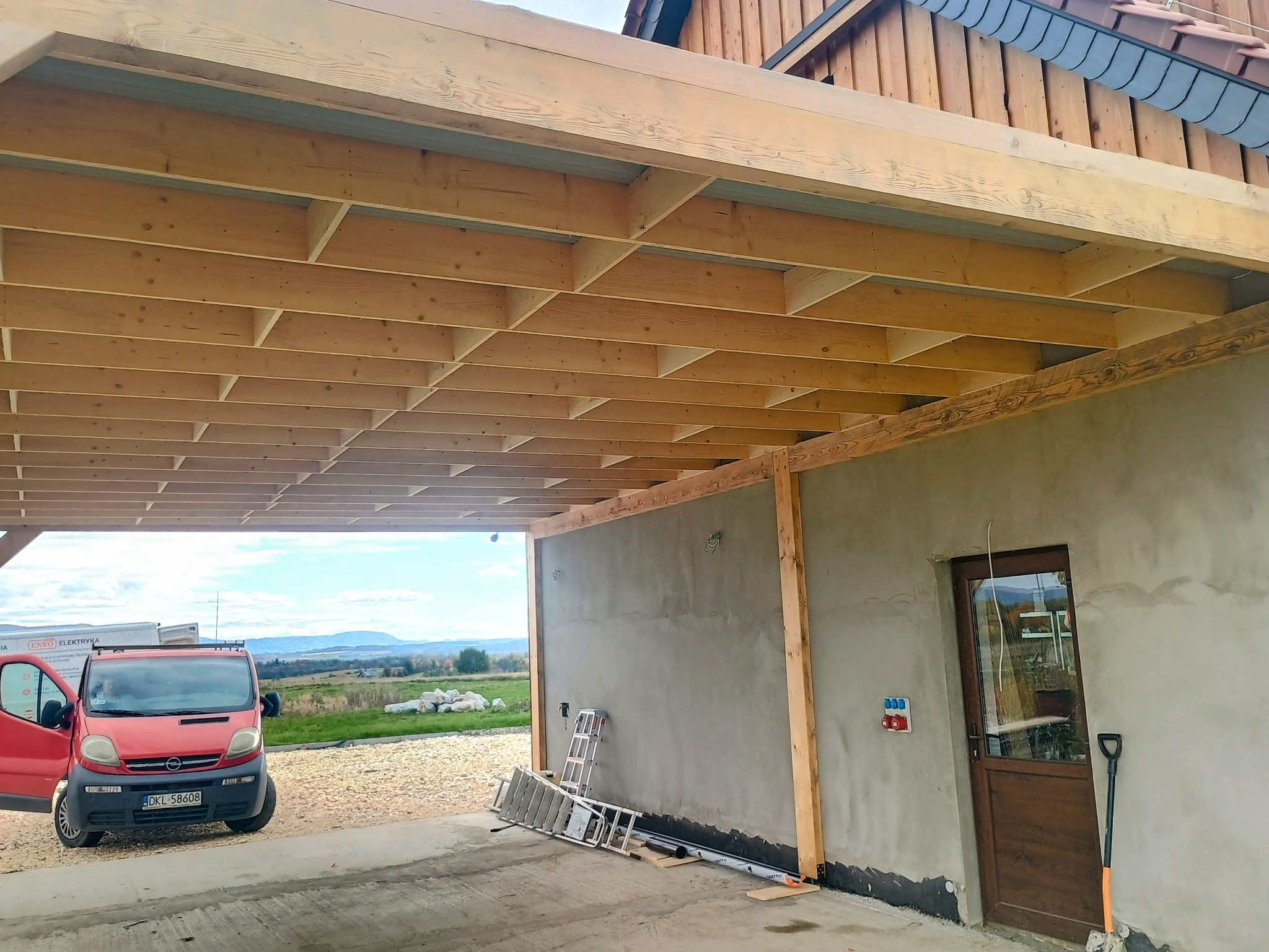 Konstrukcja z belek drewnianych tworząca zadaszenie, widok od dołu. W tle krajobraz i czerwony samochód dostawczy. Elementy budynku w stanie surowym.