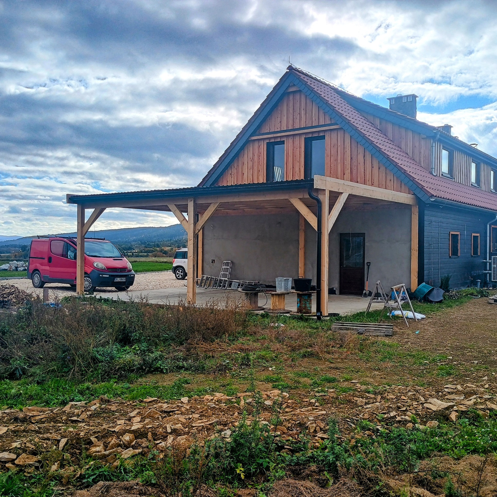 Drewniana konstrukcja zadaszenia przy domu jednorodzinnym, widok z zewnątrz. Na pierwszym planie gruz i roślinność, w tle czerwony van i górzysty krajobraz.