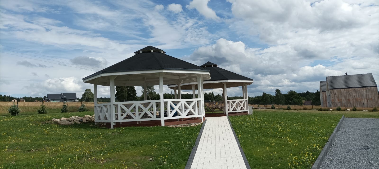 Dwie białe altany z czarnym dachem, połączone ścieżką, na tle zielonego trawnika i pochmurnego nieba. Widoczny budynek z drewnianą elewacją w oddali.