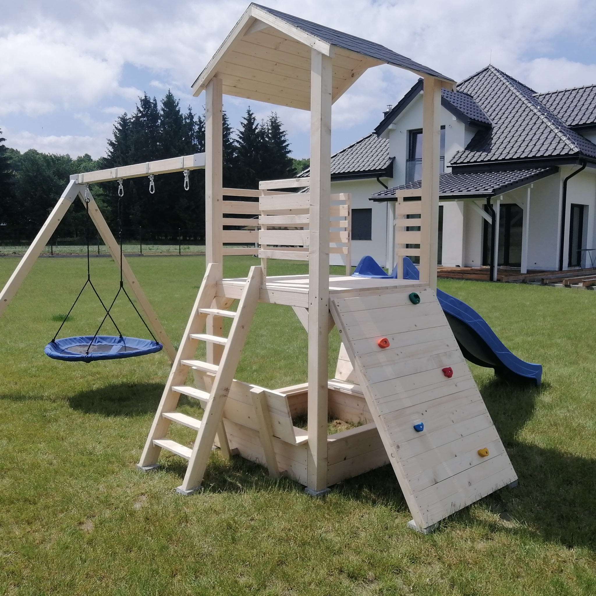 Drewniany plac zabaw z huśtawką, zjeżdżalnią i ścianką wspinaczkową na trawniku przed domem. Jasne drewno, błękitne elementy. Widok z bliska, słoneczny dzień.