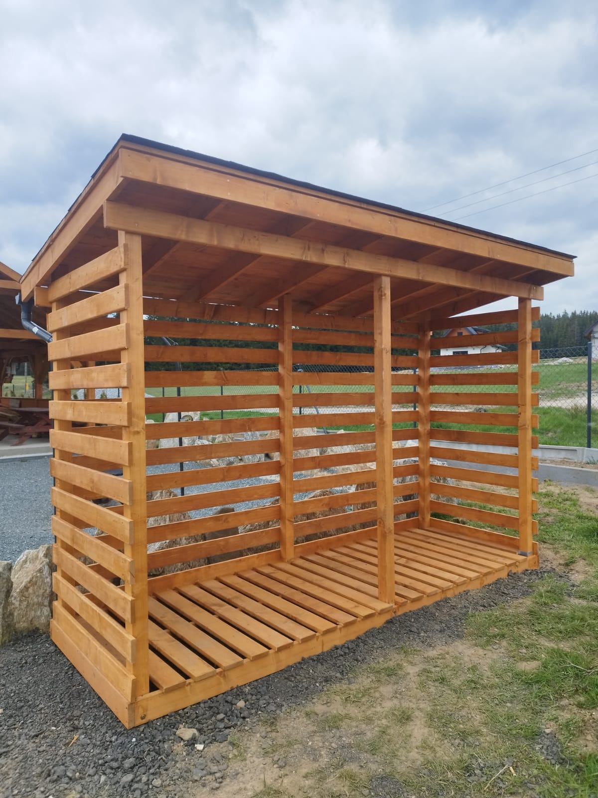 Drewniana konstrukcja z dachem, prawdopodobnie drewutnia, wykonana z jasnego drewna, z poziomymi szczeblami i podłogą z desek, na tle zieleni i pochmurnego nieba.