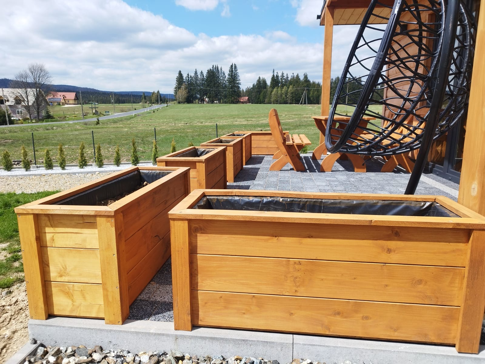 Drewniane, prostokątne donice na tarasie z szarej kostki, w tle zielony krajobraz i wiszące krzesło. Donice wyłożone czarną folią, gotowe do sadzenia roślin.