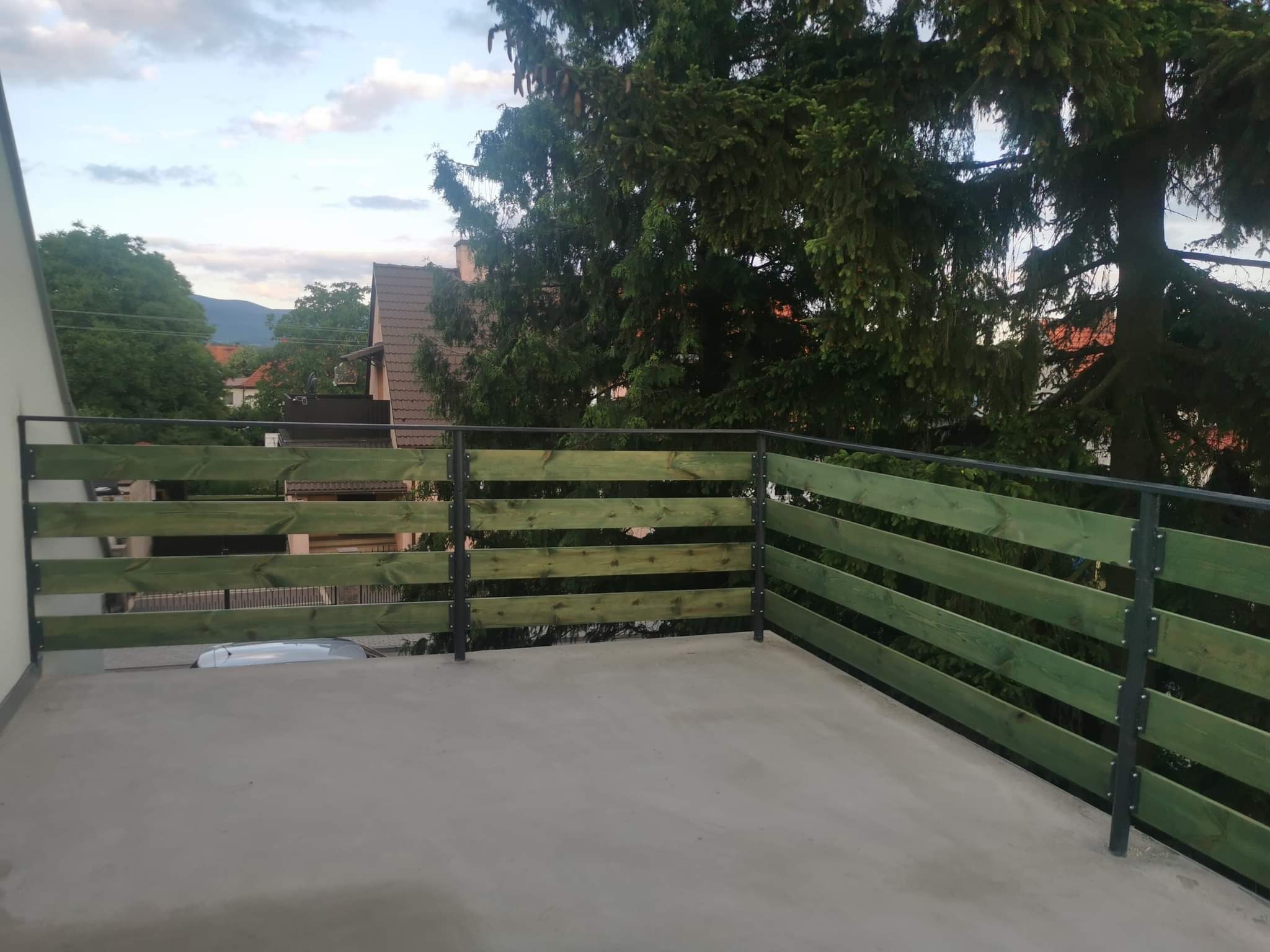 Nowoczesna, zielona balustrada drewniana z metalowymi słupkami na betonowym tarasie. W tle zieleń drzew i zabudowa mieszkalna. Ujęcie z góry.