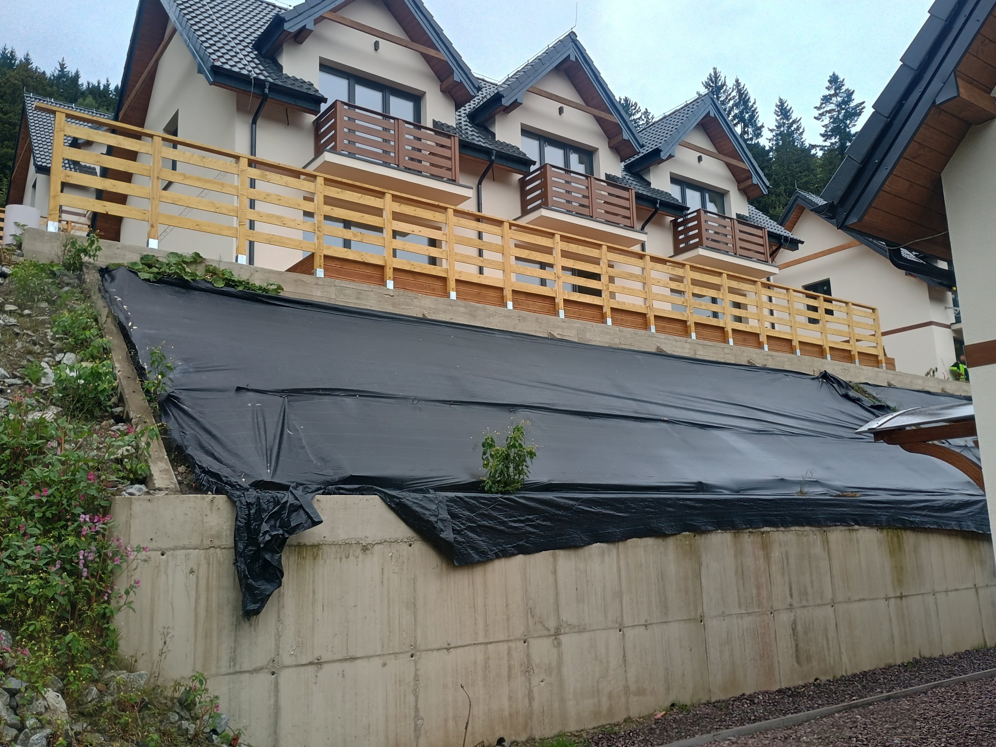 Nowoczesny dom z drewnianymi balustradami balkonowymi i ogrodzeniem tarasu. Czarna folia zabezpieczająca skarpę. Widok z dołu, pochmurne niebo.