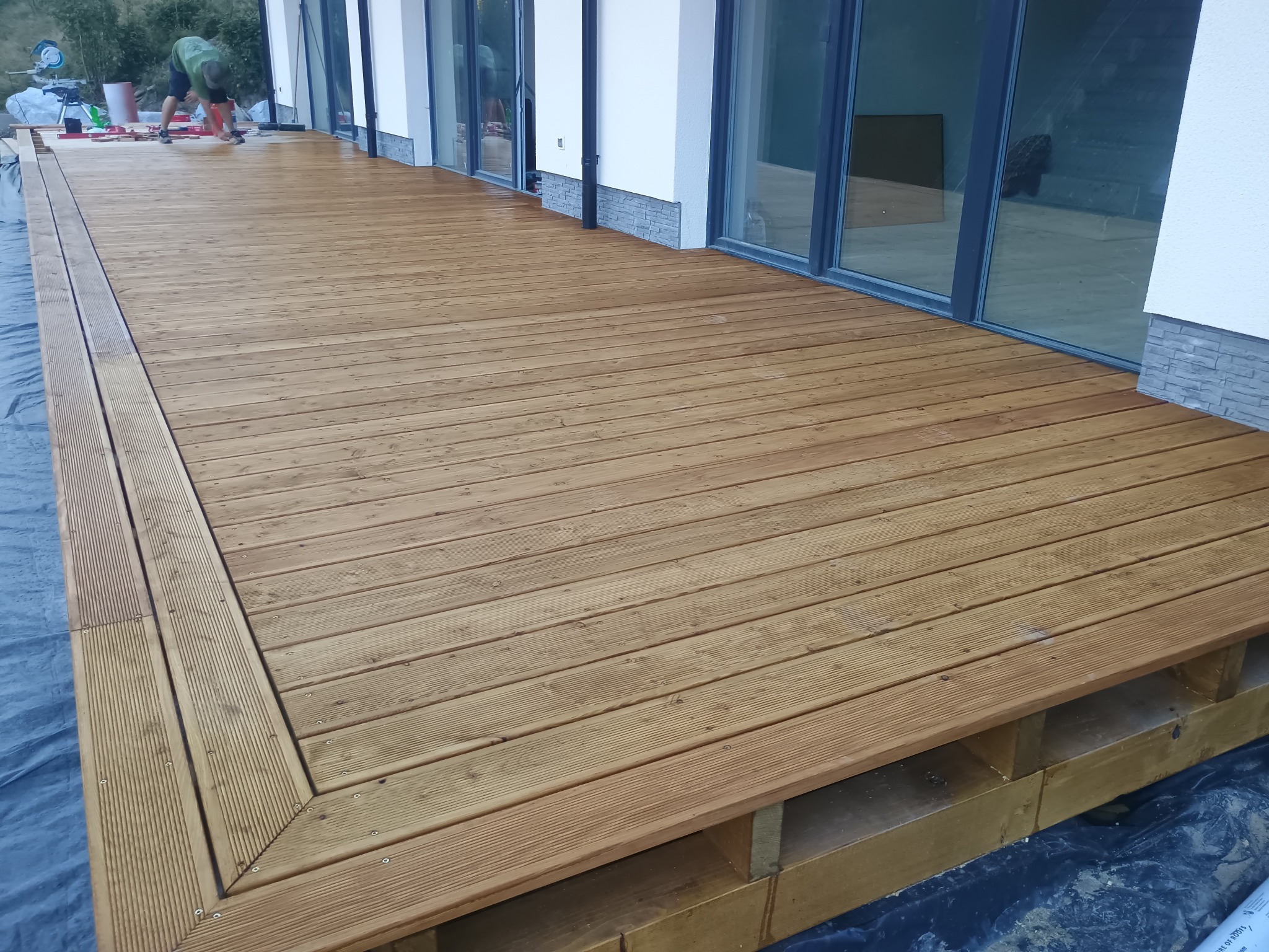 Jasny, drewniany taras z widoczną konstrukcją nośną, przylegający do budynku z dużymi oknami. Osoba w tle wykonuje prace wykończeniowe.