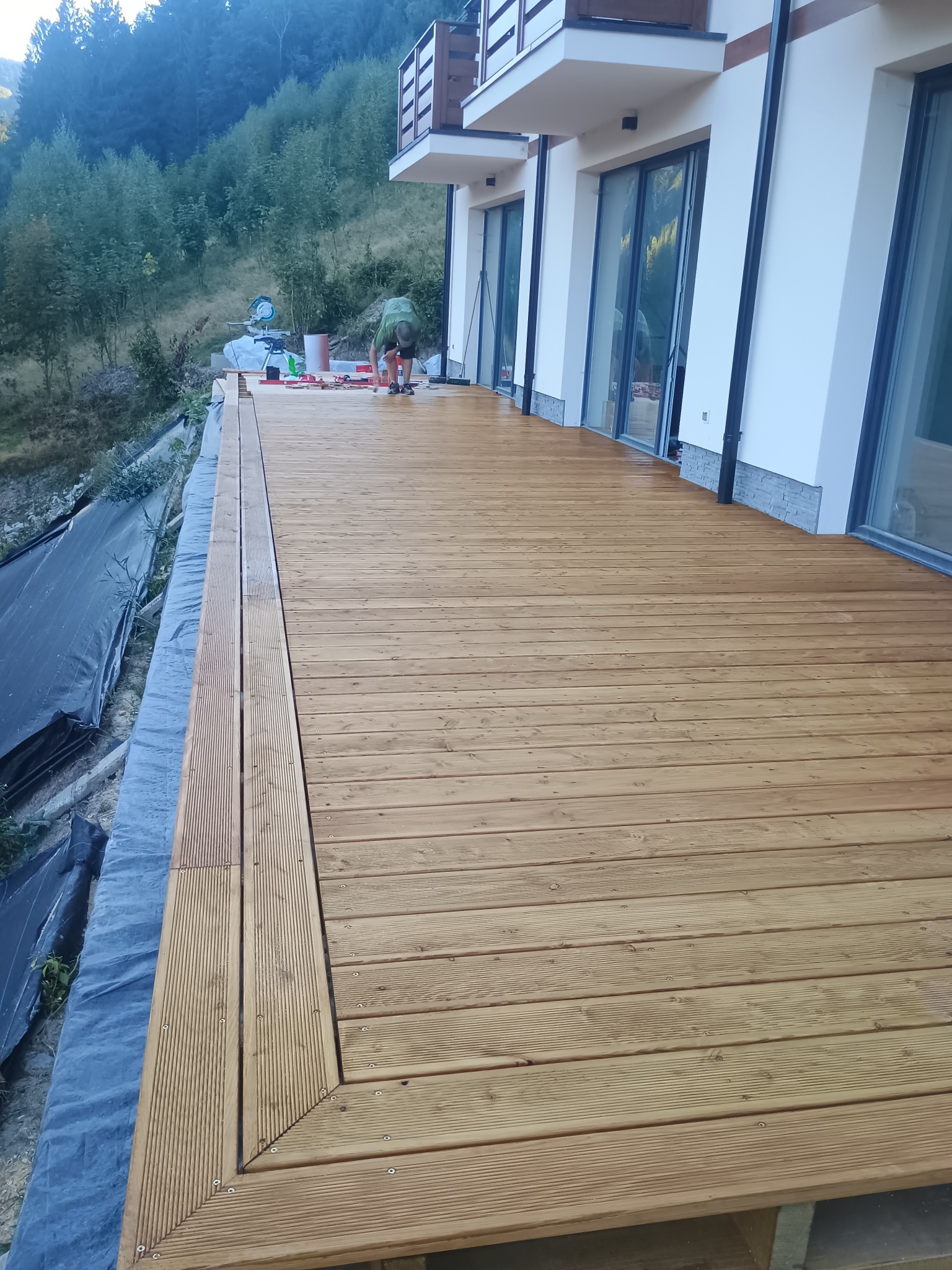 Wykończony taras z desek na zewnątrz budynku, z widocznym fragmentem pracującego stolarza i narzędziami. Deski ułożone równolegle, z estetycznym wykończeniem krawędzi.