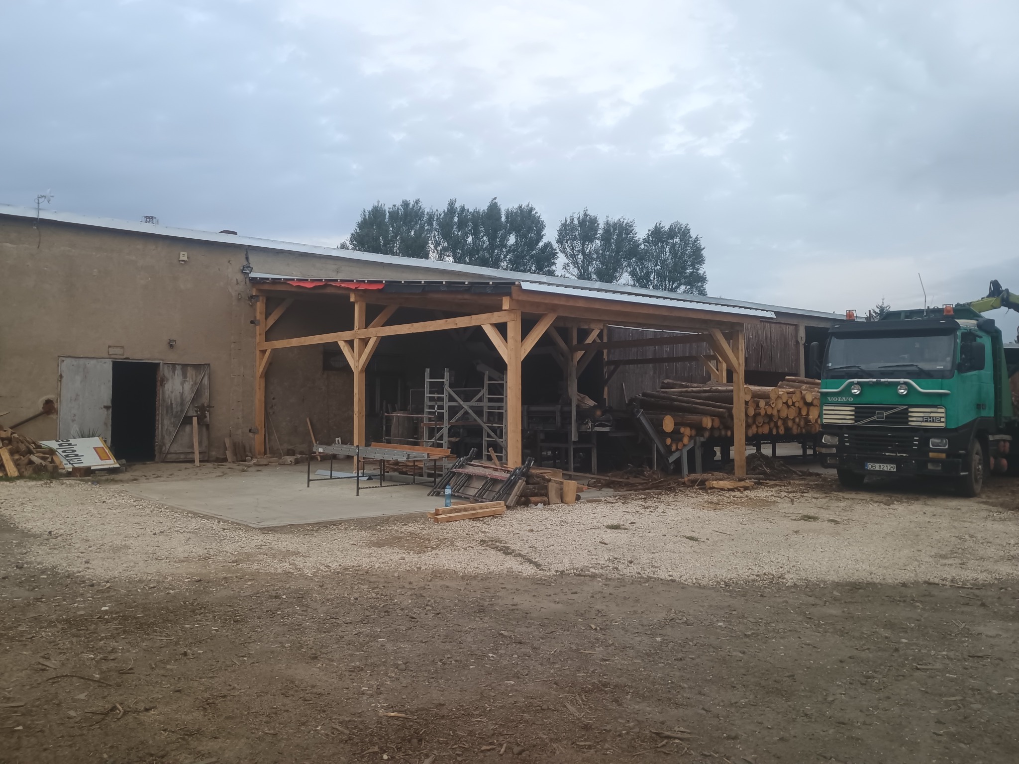 Konstrukcja z drewna w trakcie budowy, widoczny szkielet z belek, na zewnątrz stoi ciężarówka z drewnem, całość na tle budynku gospodarczego i pochmurnego nieba.