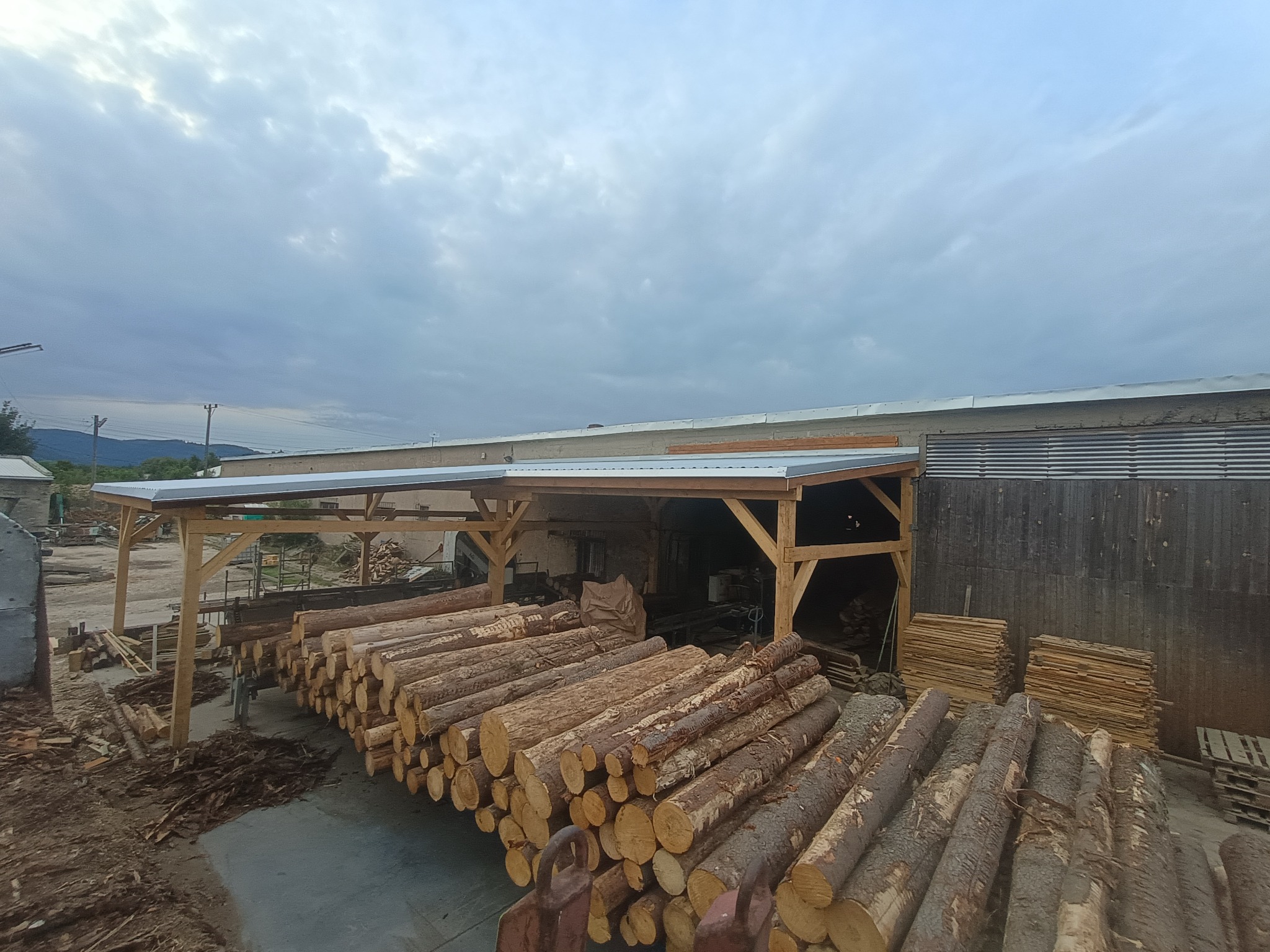 Składowane bale drewna pod zadaszeniem z drewnianą konstrukcją. W tle budynek i pochmurne niebo. Widoczne deski i elementy konstrukcyjne.