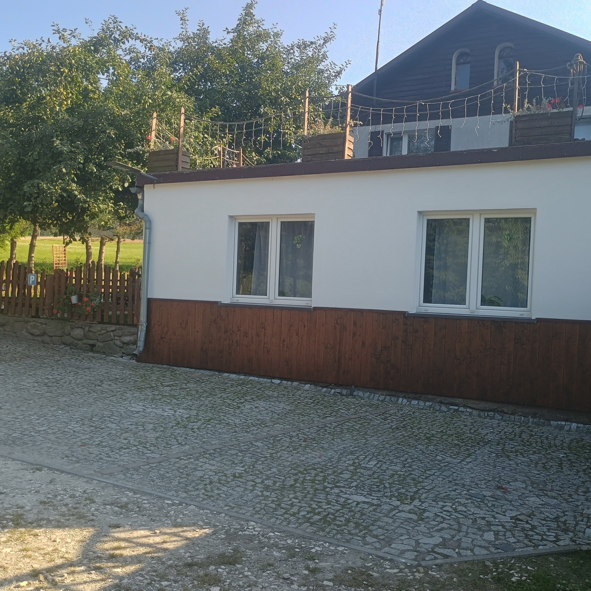 Biała ściana budynku z drewnianą boazerią i dwoma oknami, widok z zewnątrz na brukowany podjazd i drewniane ogrodzenie z roślinami. Na dachu dekoracyjna siatka.