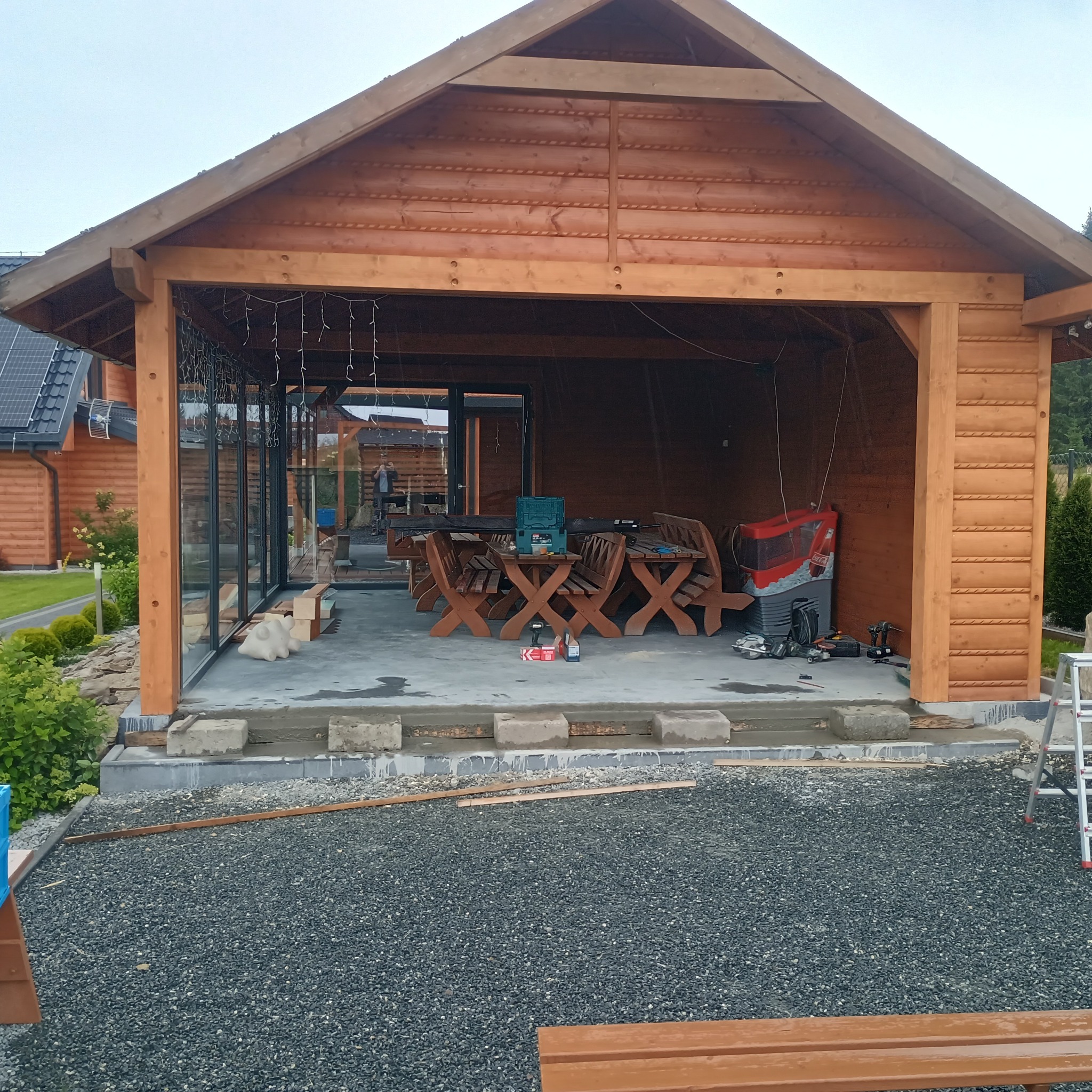 Drewniana altana w trakcie budowy, widoczne narzędzia i materiały. Częściowo przeszklona ściana, surowe wykończenie podłogi. W tle dom z panelami słonecznymi.
