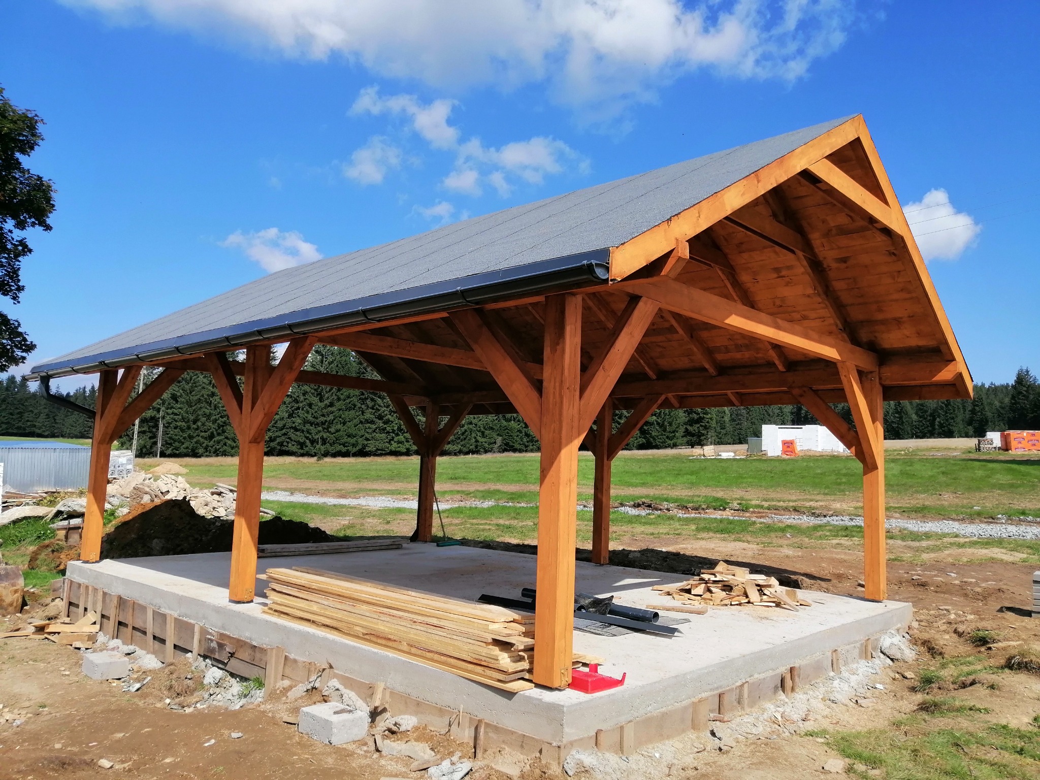 Konstrukcja drewniana z szarym dachem na betonowej podstawie, w tle zielona łąka i las, widoczne przygotowane deski i elementy instalacji.