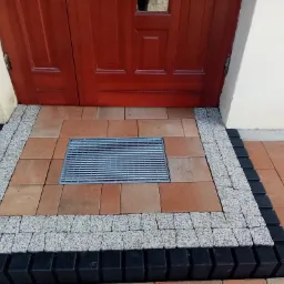 Ułożona kostka brukowa przed wejściem do domu z kratką ściekową, obramowana szarymi i czarnymi kostkami, widoczne czerwone drzwi wejściowe i fragment białej ściany.