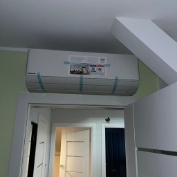 ADAPIA SPÓŁKA Z OGRANICZONĄ ODPOWIEDZIALNOŚCIĄ - Zamontowana jednostka wewnętrzna klimatyzacji Panasonic, widoczna nad drzwiami. Zabezpieczona taśmą montażową. Widok z dołu, fragment sufitu i ściany w tle.
