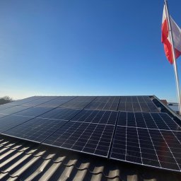 ADAPIA SPÓŁKA Z OGRANICZONĄ ODPOWIEDZIALNOŚCIĄ - Panele fotowoltaiczne na dachu z dachówki ceramicznej, w tle błękitne niebo i powiewająca flaga Polski na maszcie. Widok z dołu, słoneczny dzień.