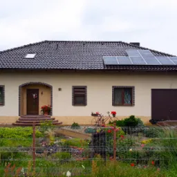 Jednorodzinny dom z brązowym dachem, na którym zamontowano panele fotowoltaiczne; w ogrodzie ozdobny rower z kwiatami.