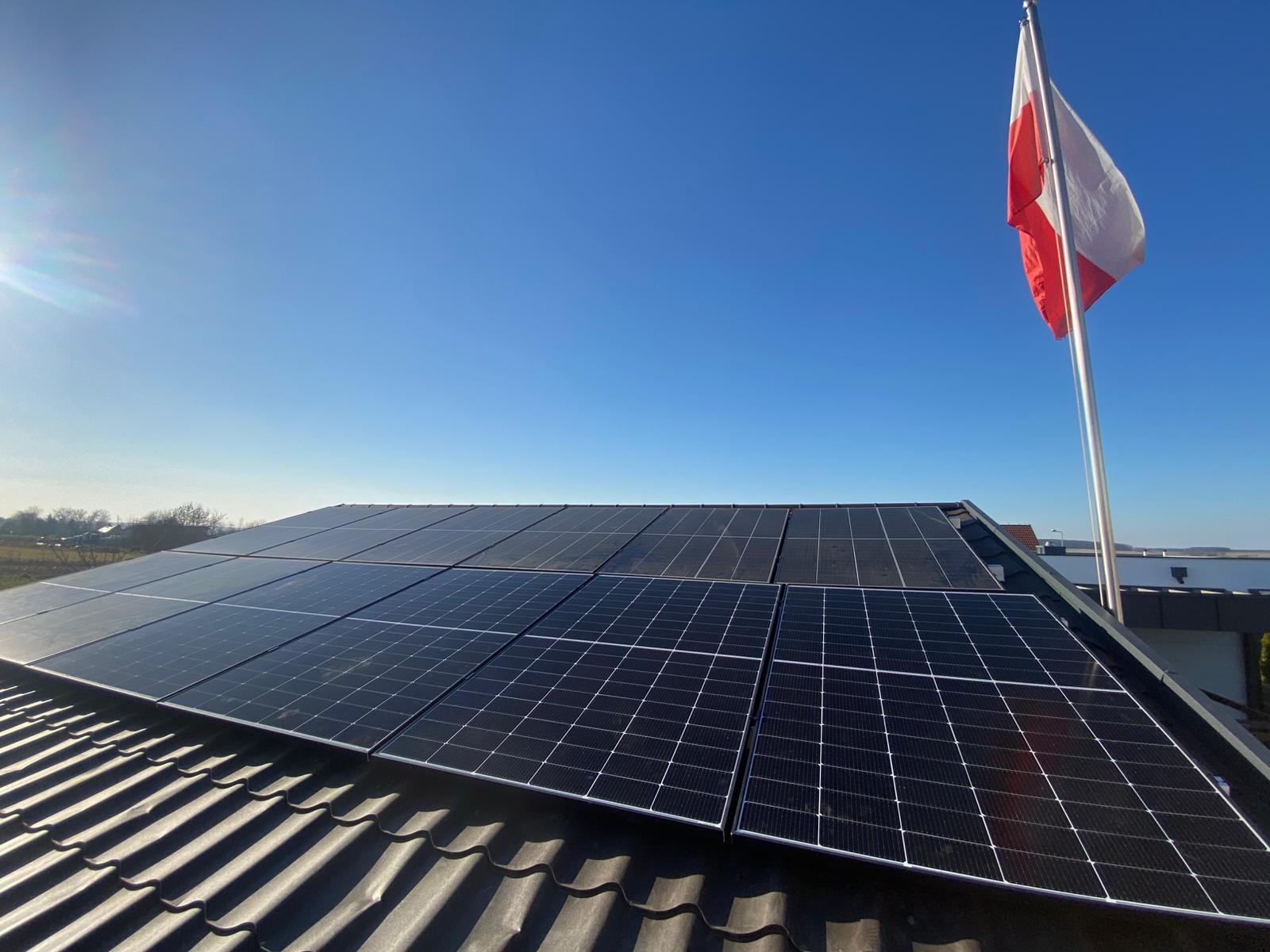 Panele fotowoltaiczne na dachu z dachówki ceramicznej, w tle błękitne niebo i powiewająca flaga Polski na maszcie. Widok z dołu, słoneczny dzień.