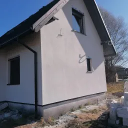 Boczny widok na dom jednorodzinny z jasną elewacją, czarną stolarką okienną i orynnowaniem, widoczne przewody elektryczne na ścianie oraz stos materiałów budowlanych na posesji.