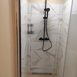 Kabina prysznicowa z czarną armaturą, wyłożona białymi płytkami z marmurkowym wzorem, uzupełniona o półkę na kosmetyki.