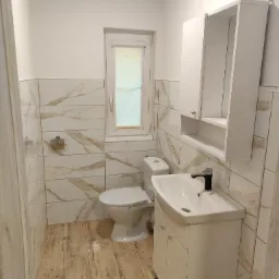 Nowoczesna, jasna łazienka z umywalką na szafce, toaletą i beżowo-białymi płytkami imitującymi marmur na ścianach, uzupełniona drewnopodobną podłogą i białymi szafkami.