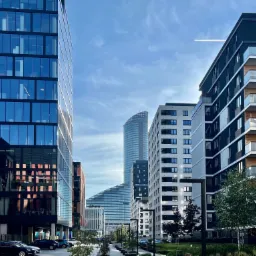 Nowoczesna architektura miejska: szklane biurowce i apartamentowce w słoneczny dzień, z widocznym śladem samolotu na niebie.