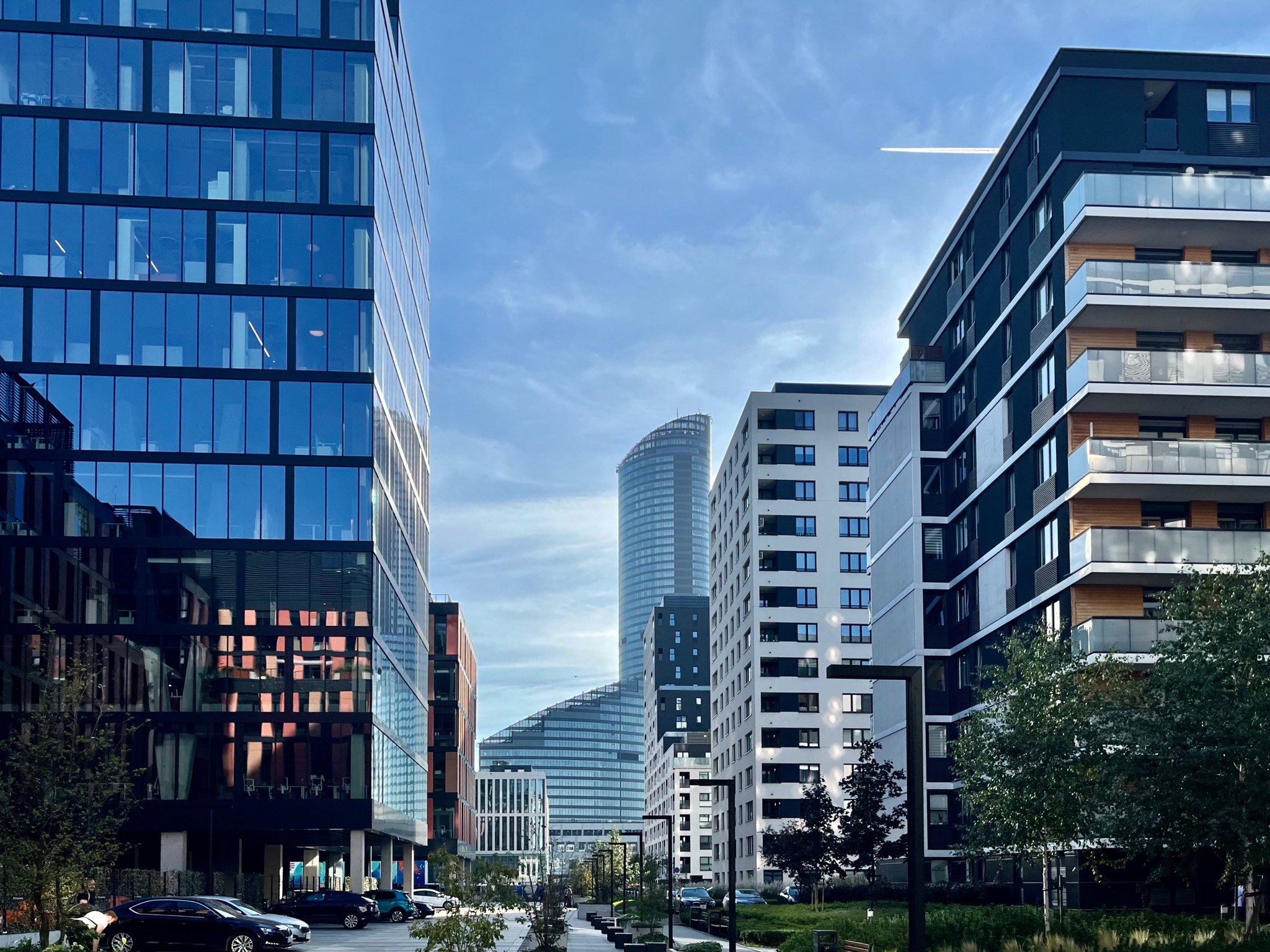 Nowoczesna architektura miejska: szklane biurowce i apartamentowce w słoneczny dzień, z widocznym śladem samolotu na niebie.