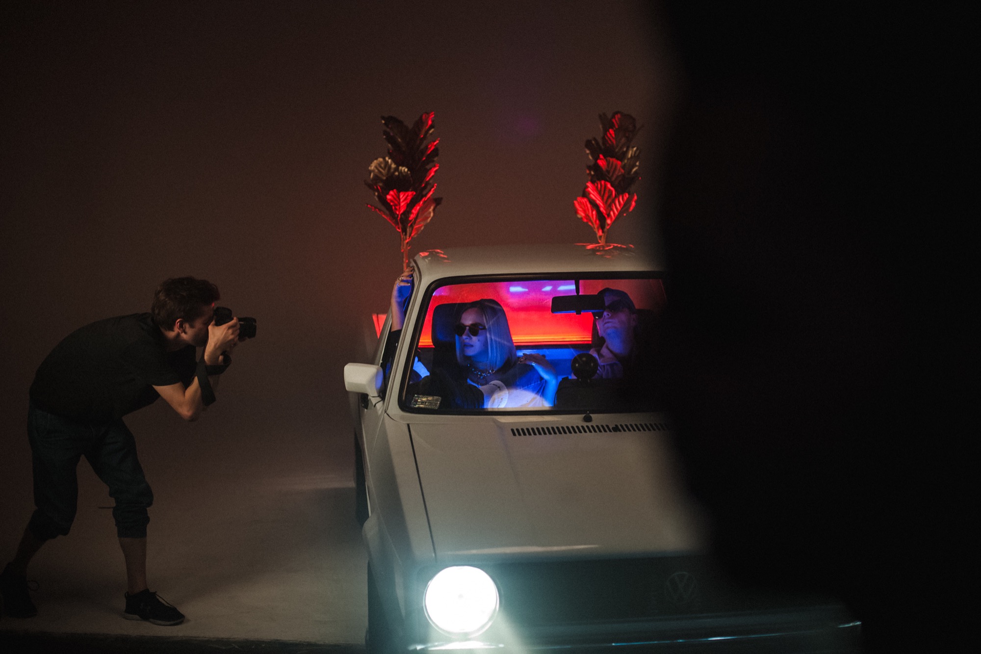 Fotograf robi zdjęcie dwóm osobom w białym samochodzie VW Golf z rekwizytami w postaci roślin na dachu, oświetlonymi czerwonym i niebieskim światłem.