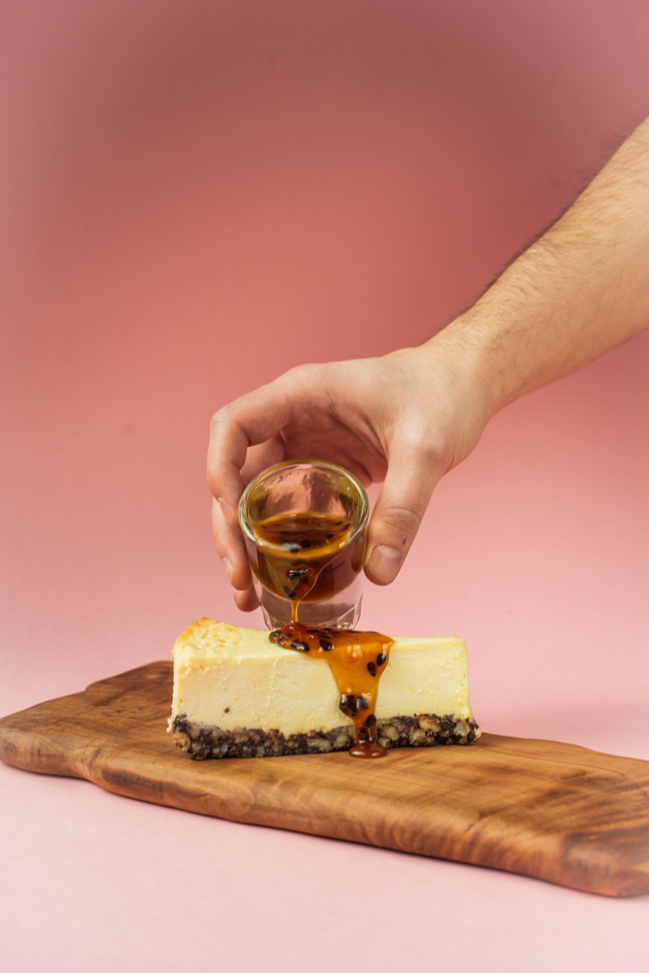 Kawałek sernika na desce drewnianej, polewany sosem marakuja z kieliszka przez męską dłoń na różowym tle.