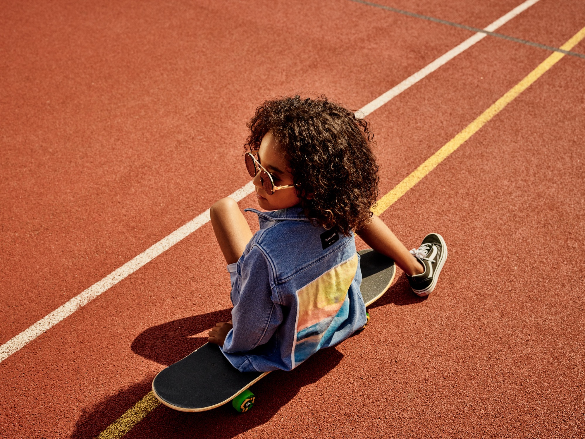 Młoda dziewczyna z kręconymi włosami i okularach przeciwsłonecznych siedzi na deskorolce na czerwonej bieżni z białymi i żółtymi liniami, ubrana w jeansową kurtkę z kolorowym nadrukiem na plecach.