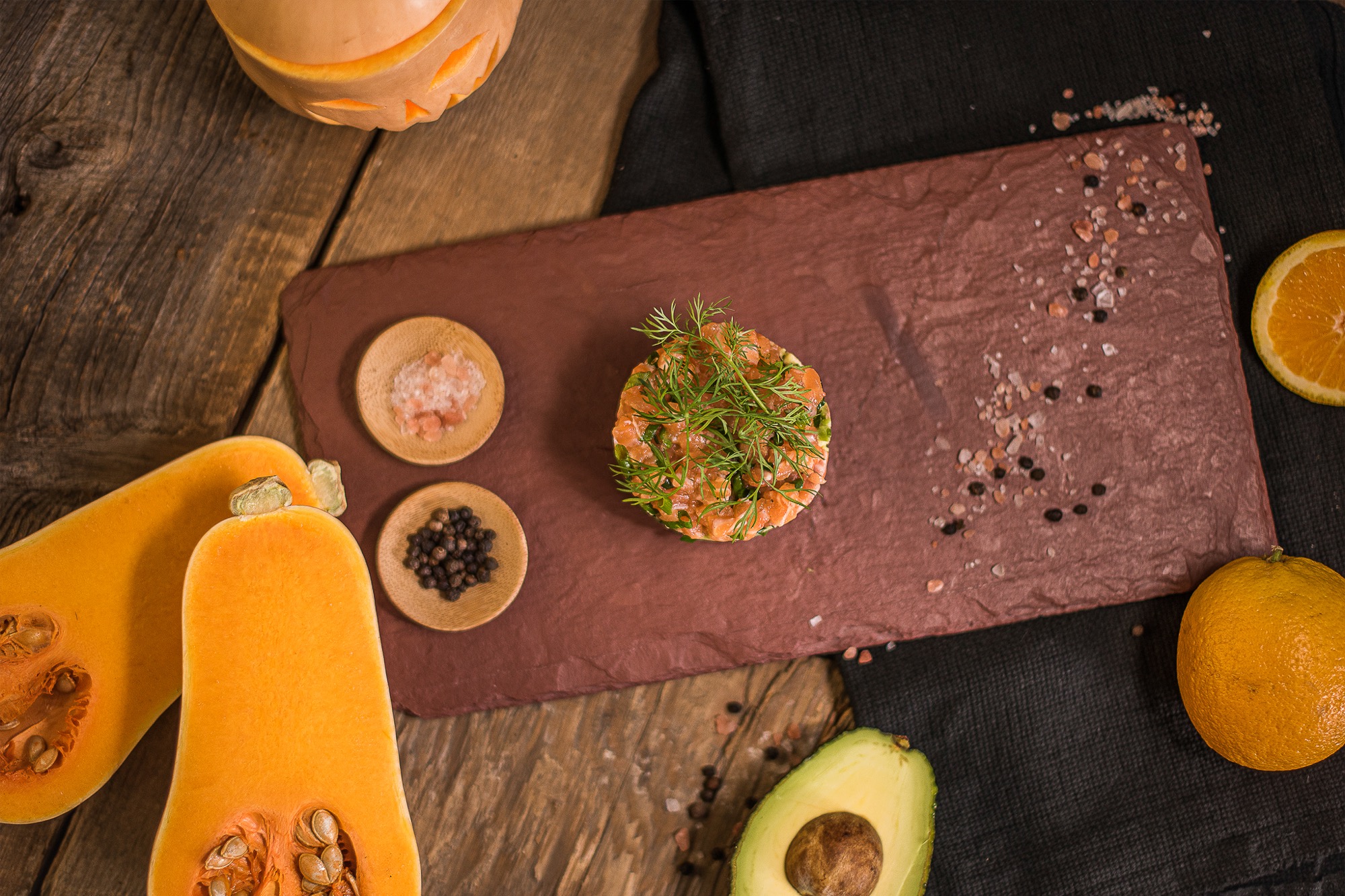Tartar z łososia udekorowany koperkiem na kamiennej desce, otoczony dynią, awokado, pomarańczą i przyprawami na drewnianym stole.