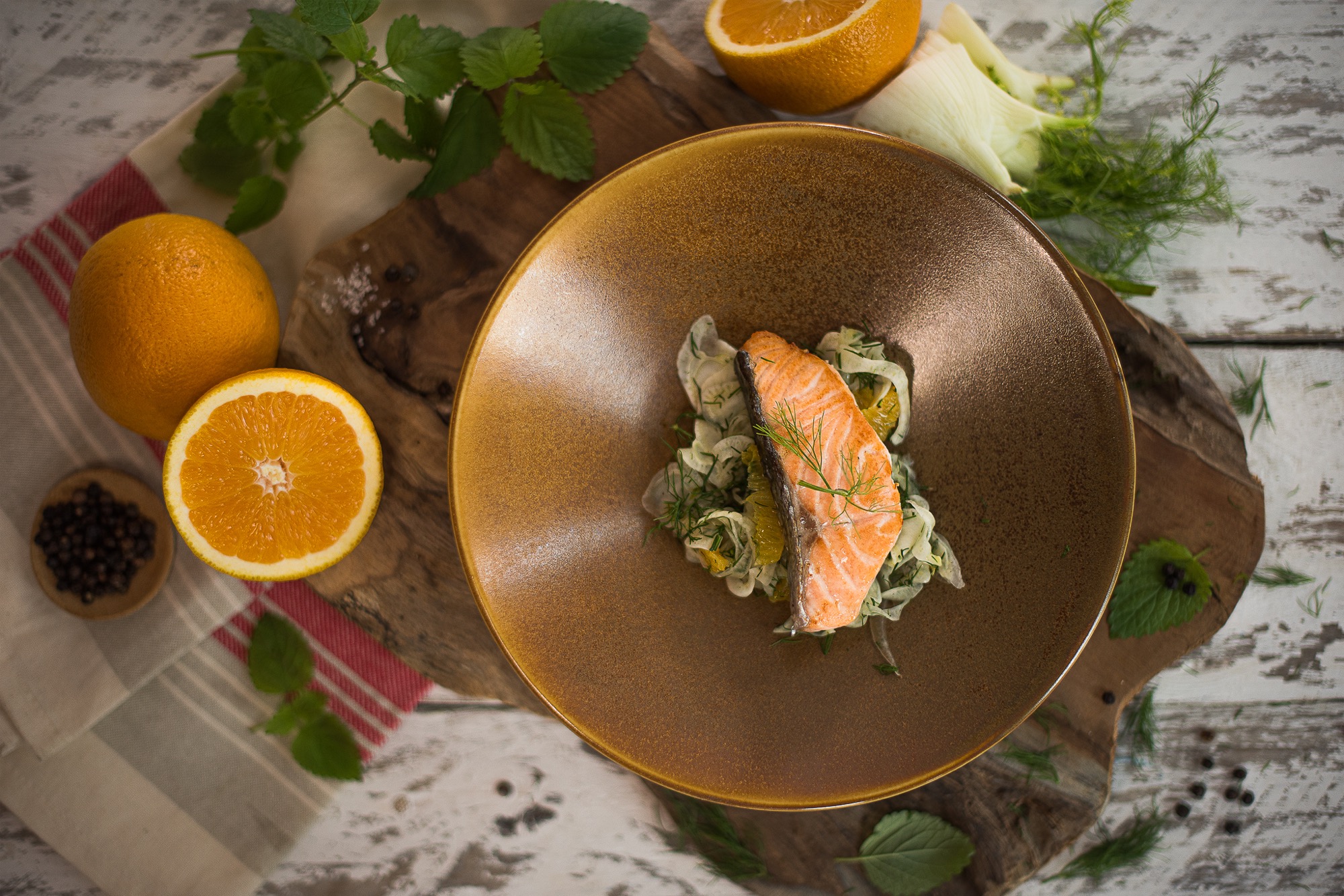 Filet z łososia na brązowym talerzu z cytrusami i fenkułem, otoczony pomarańczami, pieprzem i ziołami na drewnianym blacie.