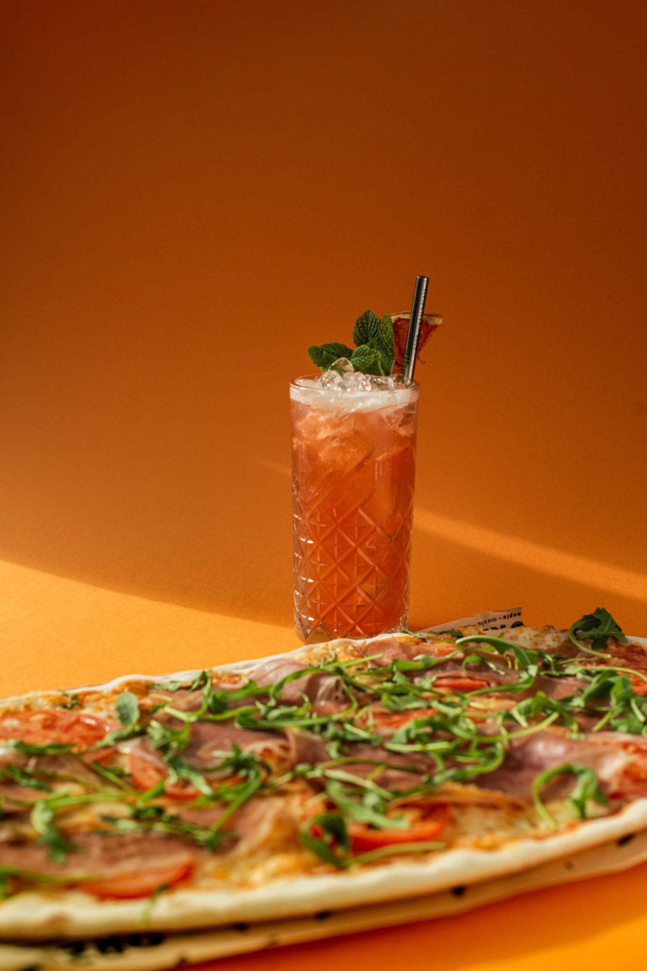 Pomarańczowy koktajl z lodem, miętą i suszoną pomarańczą w szklance o szlifowanej fakturze, obok długa pizza z szynką parmeńską, pomidorami i rukolą na pomarańczowym tle.