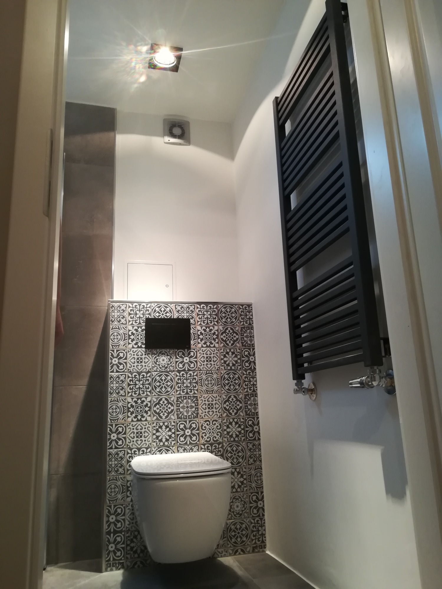 Nowoczesna toaleta z czarno-białymi płytkami dekoracyjnymi, wiszącą miską WC i czarnym grzejnikiem drabinkowym, oświetlona punktowo