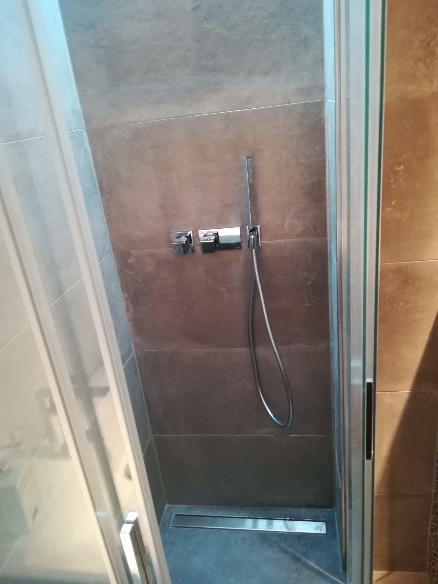 Kabina prysznicowa z szarymi płytkami, chromowaną armaturą i odpływem liniowym w podłodze, widok z zewnątrz przez częściowo otwarte szklane drzwi.