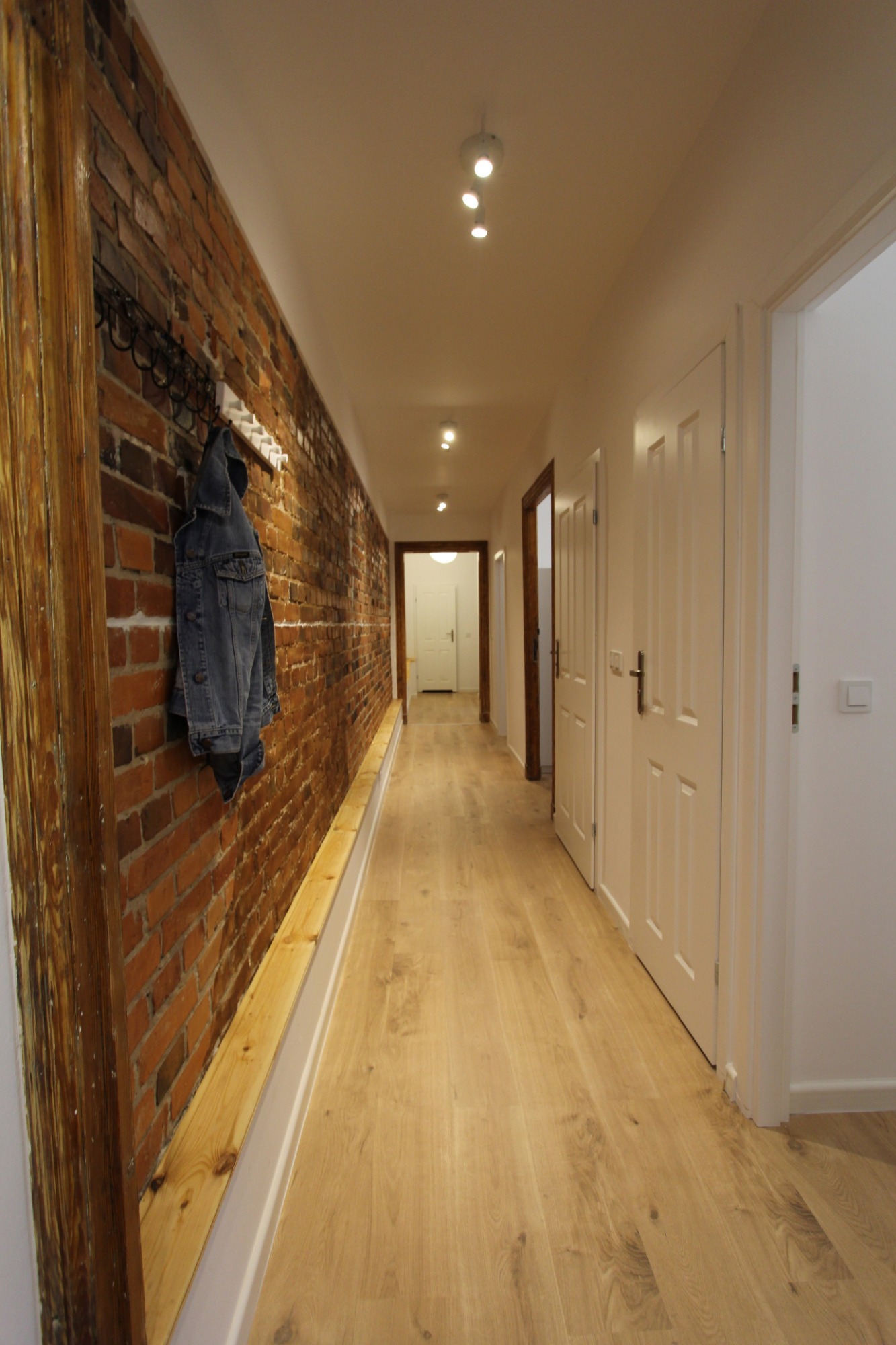 Długi korytarz po renowacji z ceglaną ścianą, drewnianą boazerią, jasnymi panelami podłogowymi i białymi drzwiami; dżinsowa kurtka wisząca na wieszaku.