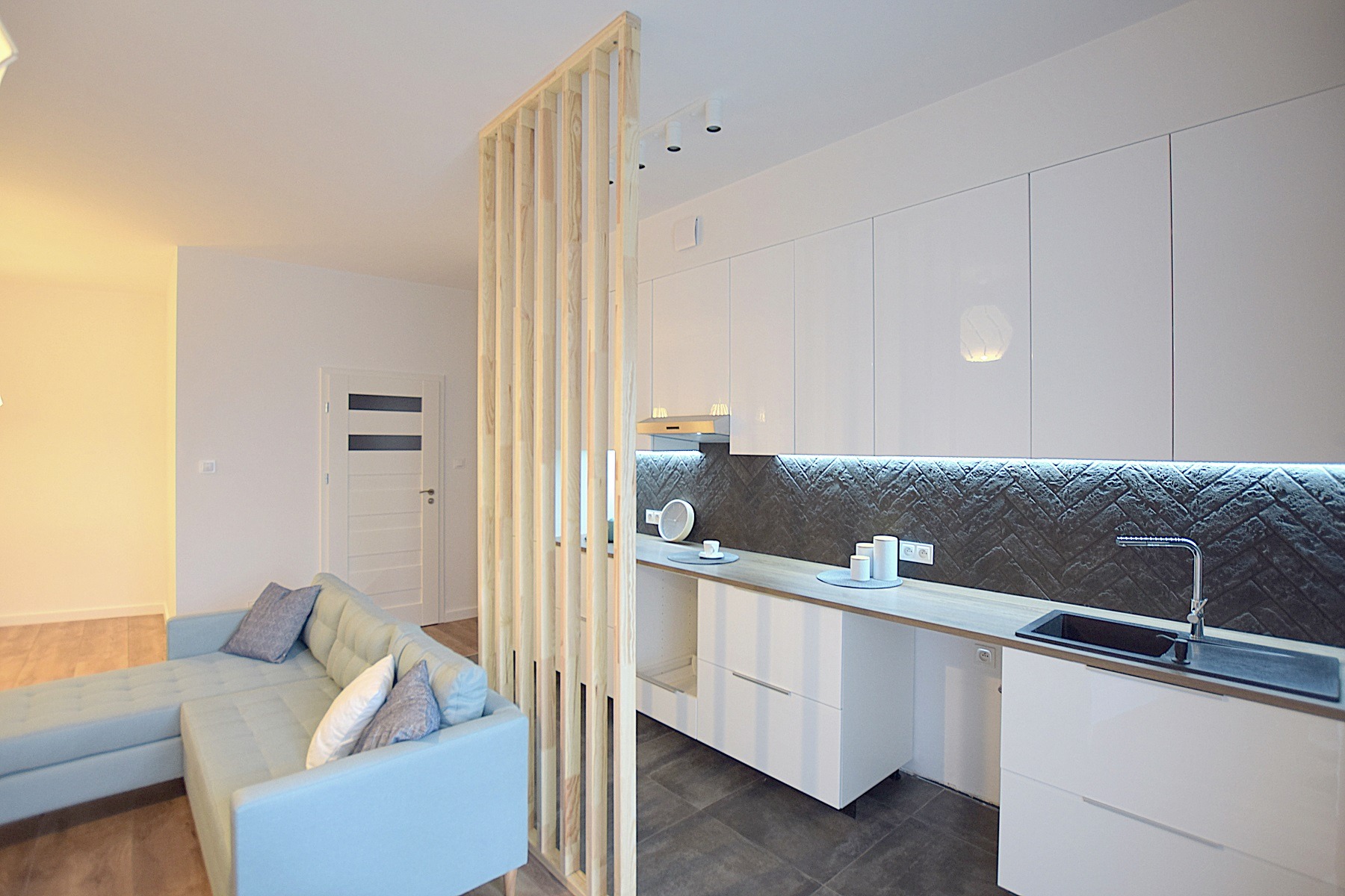 Nowoczesna kuchnia z białymi błyszczącymi szafkami i ciemną, wzorzystą ścianą nad blatem, oddzielona od salonu drewnianą ażurową ścianką działową, widoczny fragment turkusowej sofy.