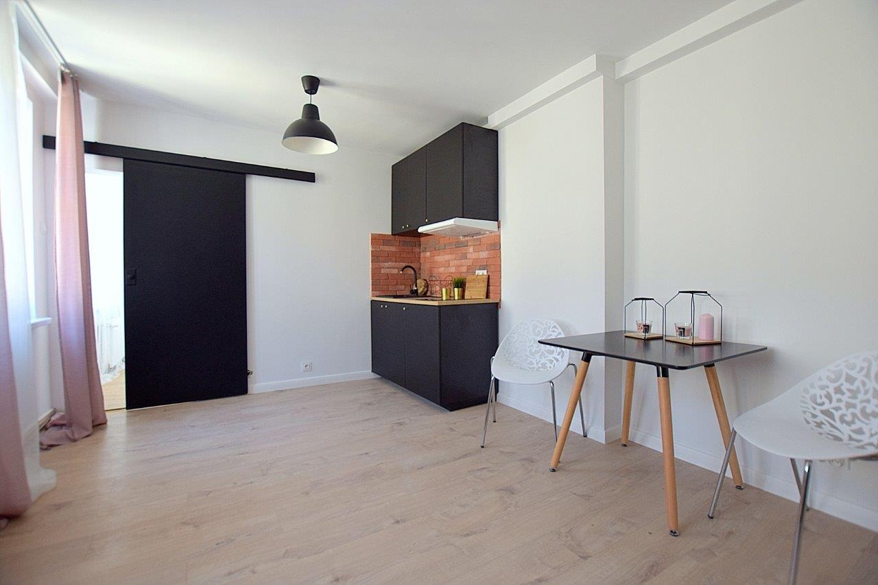 Jasne wnętrze mieszkania z aneksem kuchennym w ciemnej zabudowie, ceglaną ścianą nad blatem, przesuwnymi drzwiami w kolorze czarnym i drewnianą podłogą; stół z czarnym blatem i białymi krzesłami.