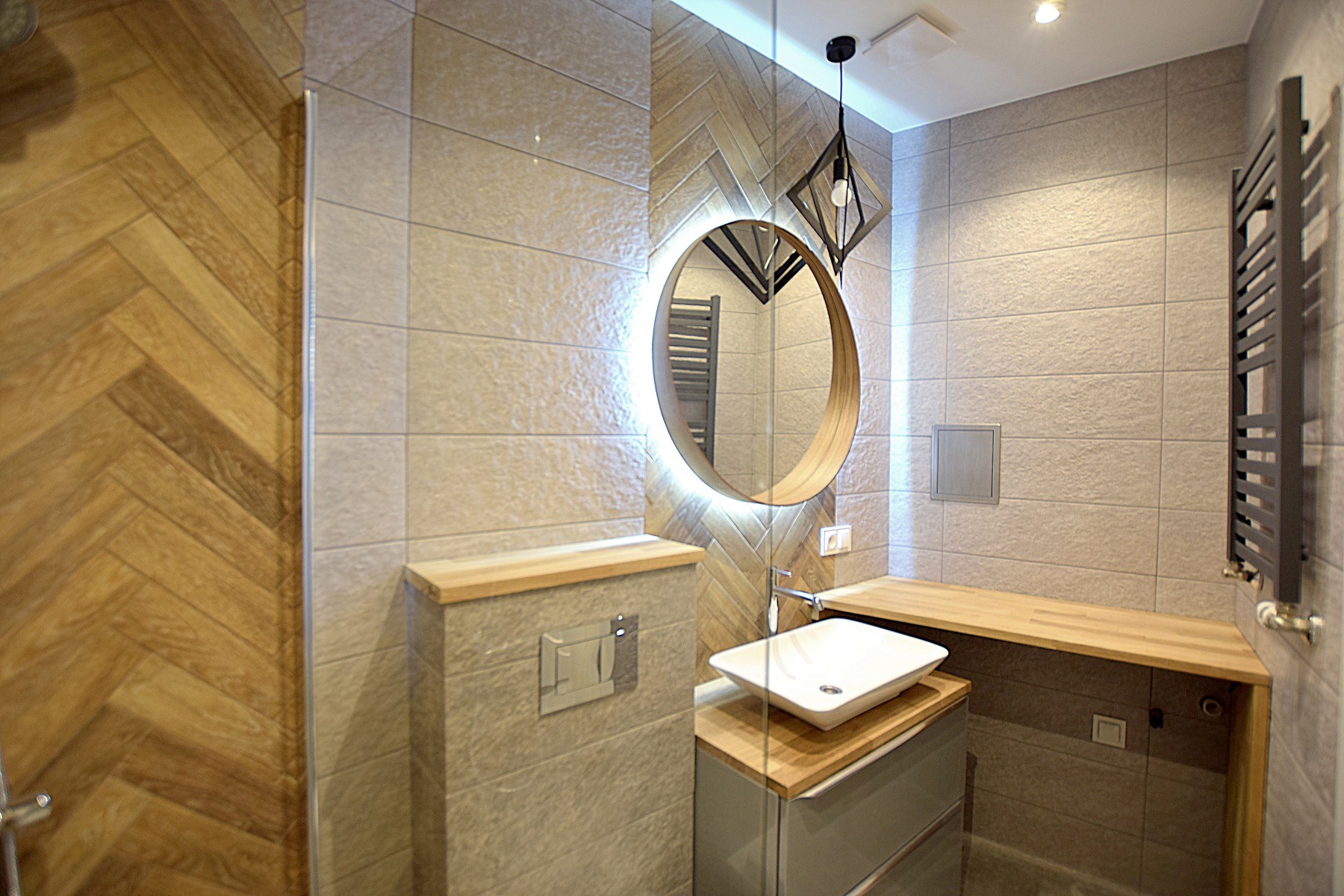 Nowoczesna łazienka z płytkami imitującymi drewno ułożonymi w jodełkę na ścianie kabiny prysznicowej, okrągłe lustro z podświetleniem LED, biała umywalka nablatowa na szafce z drewnianym blatem...
