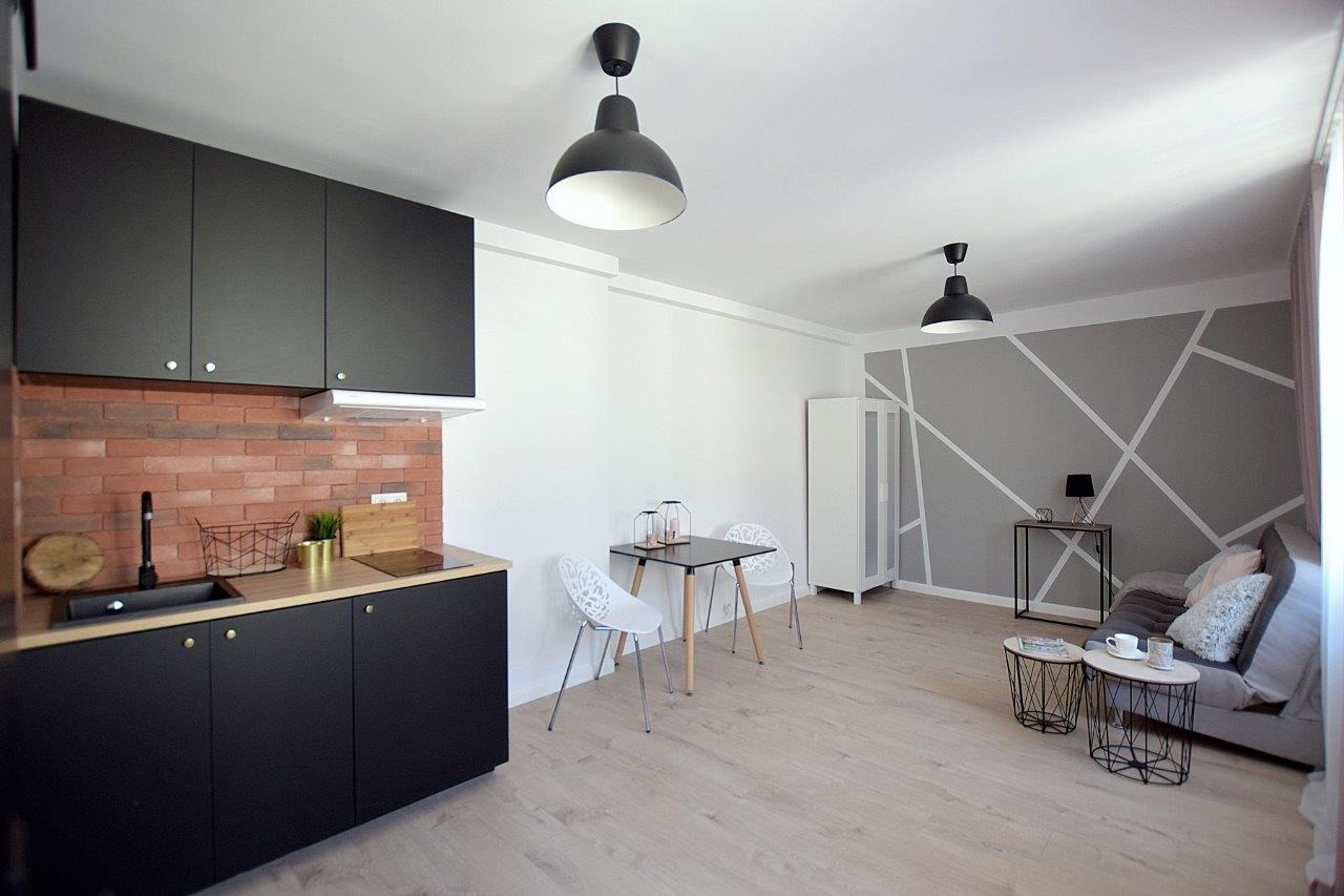 Nowoczesna aranżacja wnętrza kawalerki z aneksem kuchennym: czarne szafki kuchenne z ceglaną ścianą, drewniany blat, stół z krzesłami, szara ściana z geometrycznym wzorem, drewniana podłoga...
