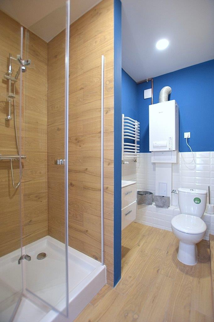 Nowoczesna łazienka z kabiną prysznicową typu walk-in, drewnopodobnymi płytkami na ścianach i podłodze, niebieską ścianą oraz białym bojlerem gazowym i grzejnikiem drabinkowym.