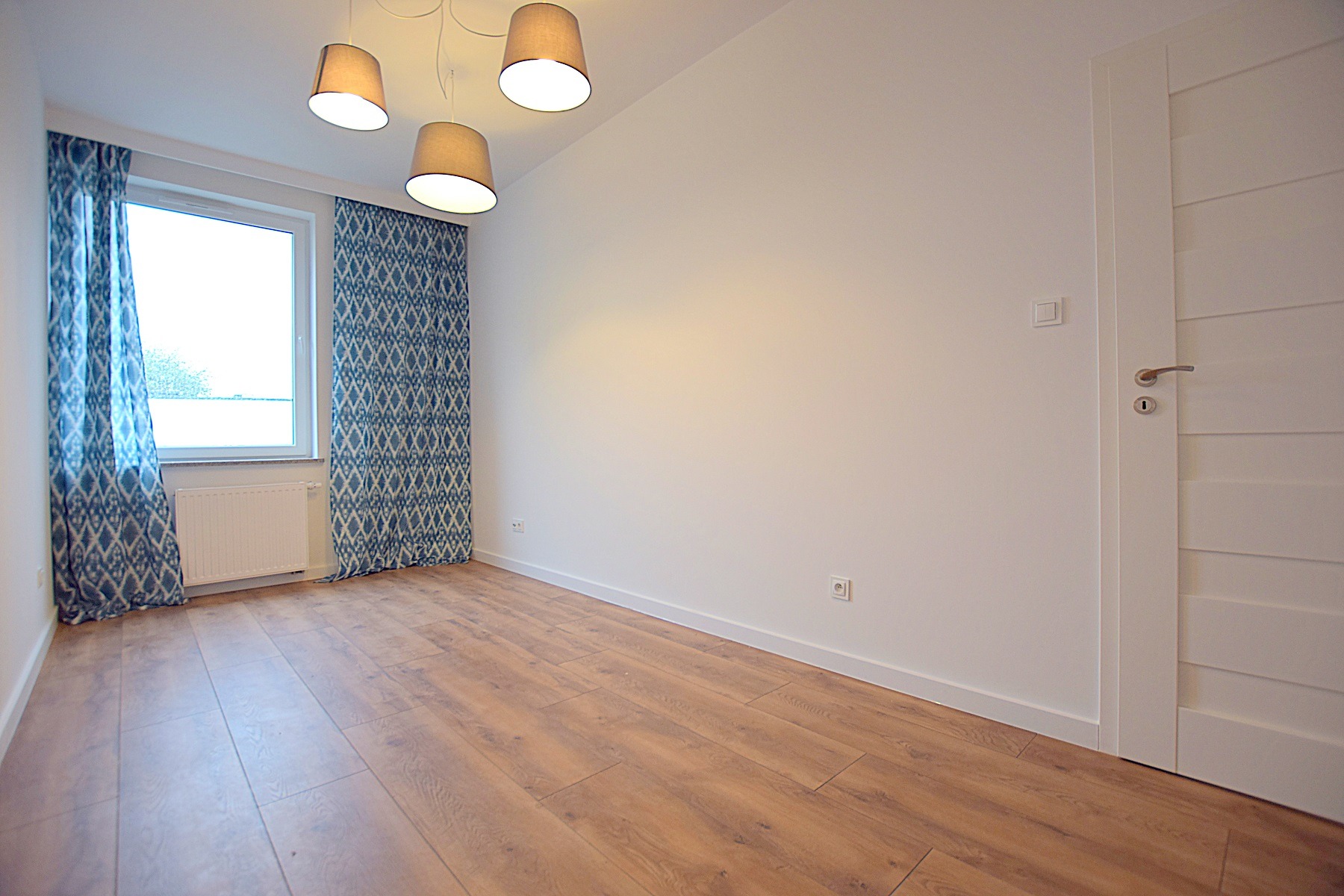 Pusty pokój z panelami podłogowymi w odcieniach brązu, białymi ścianami, grzejnikiem pod oknem z niebieskimi zasłonami w geometryczne wzory oraz trzema lampami z beżowymi abażurami.