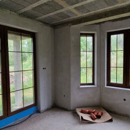 LUKMAR SPÓŁKA Z OGRANICZONĄ ODPOWIEDZIALNOŚCIĄ - Wnętrze w stanie deweloperskim z trzema oknami PCV w kolorze ciemnego drewna, widok na zewnątrz na zieleń, na podłodze karton z rurami kanalizacyjnymi.