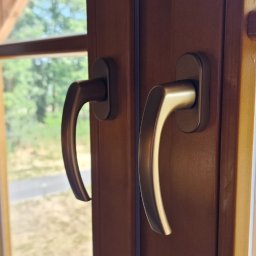 LUKMAR SPÓŁKA Z OGRANICZONĄ ODPOWIEDZIALNOŚCIĄ - Brązowe okno PCV z dwiema klamkami, widok na zewnątrz z zielenią i budynkiem w tle, naklejka producenta w rogu szyby.