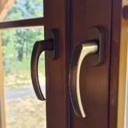 Brązowe okno PCV z dwiema klamkami, widok na zewnątrz z zielenią i budynkiem w tle, naklejka producenta w rogu szyby.