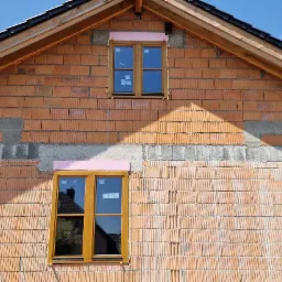 Surowy stan budynku z czerwonej cegły z zamontowanymi oknami z ramami w kolorze drewna, zabezpieczonymi różową pianką montażową, widoczne odbicie w szybach okiennych.